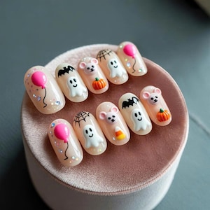 May include: A set of ten artificial nails with a Halloween theme. Each nail is painted a nude color and decorated with 3D designs including ghosts, bats, pumpkins, mice, balloons, and spiderwebs. Some nails have glitter accents.
