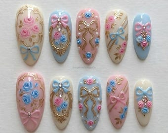 May include: A set of ten oval-shaped artificial nails. Each nail features a different design, including pink and blue roses, bows, and gold accents. Some nails have pearl embellishments. The nails are painted in pastel colors.