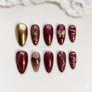 Könnte beinhalten: Ein Set von zehn künstlichen Nägeln in Gold- und Burgundertönen. Designs umfassen einen goldenen Schmetterling, ein Herz und geometrische Muster. Einige Nägel haben ein Glitzer-Finish. Maße in Zentimetern.