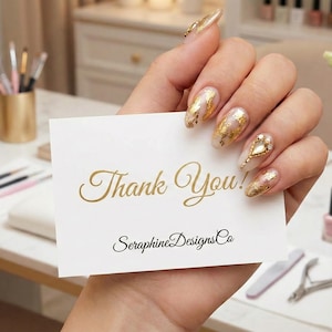 Op de afbeelding: Een hand die een witte kaart vasthoudt met gouden tekst "Thank You!" en "SeraphineDesignsCo". De nagels zijn versierd met bladgoudontwerpen. De achtergrond bevat nagellak, make-up borstels en bloemen.