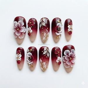 May include: A set of ten artificial nails with a gradient design, transitioning from a nude base to a deep burgundy. Each nail features 3D floral embellishments in white, burgundy, and gold, with the text "SeraphineDesignsCo" visible.