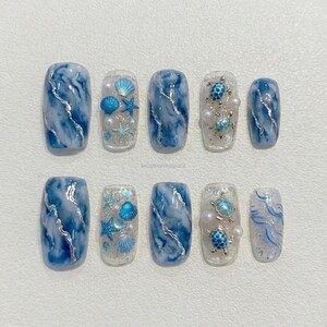 May include: A set of ten artificial nails with a sea-themed design. The nails feature a marble effect in shades of blue and white, with some adorned with seashells, starfish, and pearls. The design includes a turtle and the text "seraphinedesigns4u".