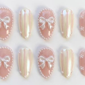 May include: A set of ten artificial nails in various designs. Some nails are light pink with a white bow and pearl border. Other nails are light pink with vertical iridescent stripes. One nail has a snowflake design.