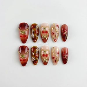 Peut inclure: Dix faux ongles en forme d'amande. Chaque ongle présente un design unique, avec des cerises, des fleurs et des chaînes dorées. La palette de couleurs comprend des nuances de rouge, de marron et de rose, avec une finition brillante. Seraphine DesignsCo est imprimé.