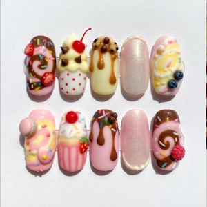 May include: A set of ten artificial nails with a dessert theme. Designs include chocolate swirls, cherries, sprinkles, and a macaron. The nails are painted in shades of pink, white, and brown, with a glossy finish.