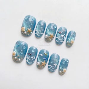 Op de afbeelding: Een set van tien blauwe kunstnagels met een glinsterende, glitterende afwerking. Elke nagel heeft ontwerpen met oceaanthema, waaronder schelpen, zeesterren, parels en kleine vissen. Sommige nagels hebben waterdruppelaccenten en witte golfpatronen.