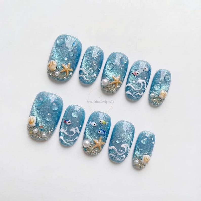 Op de afbeelding: Een set van tien blauwe kunstnagels met een glinsterende, glitterende afwerking. Elke nagel heeft ontwerpen met oceaanthema, waaronder schelpen, zeesterren, parels en kleine vissen. Sommige nagels hebben waterdruppelaccenten en witte golfpatronen.