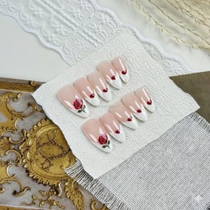 Può includere: Un set di unghie finte con punta bianca e base rosa. Alcune unghie presentano un piccolo cuore rosso, mentre altre mostrano un disegno di rosa rossa con foglie verdi. Le unghie sono disposte su una superficie bianca strutturata.
