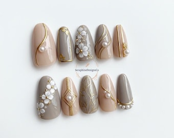 May include: A set of ten artificial nails in various designs. The nails feature a combination of neutral tones, gold accents, and pearl embellishments. Some nails have floral patterns, while others showcase geometric designs.