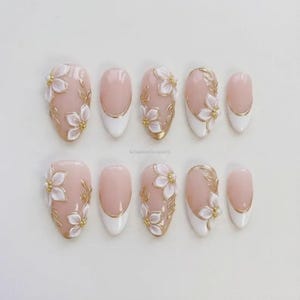 Op de afbeelding: Een set van tien kunstnagels met een lichtroze basis, witte uiteinden en gouden bloemaccenten. De nagels hebben verschillende ontwerpen, waaronder volledige bloempatronen en gouden blad details. De nagels zijn ovaal van vorm.