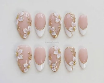 Può includere: Un set di dieci unghie finte con base rosa chiaro, punte bianche e dettagli floreali dorati. Le unghie presentano una varietà di disegni, tra cui motivi floreali completi e dettagli di foglie dorate. Le unghie sono ovali.