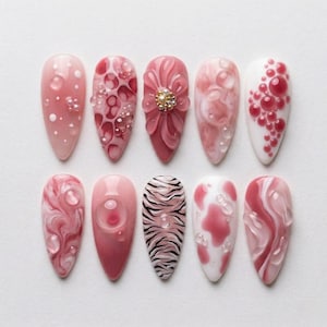 Puede incluir: Una colección de diez uñas artificiales en forma de almendra con varios diseños. Las uñas presentan tonos de rosa, blanco y transparente, con adornos como burbujas, motivos florales y un estampado de cebra. Algunas uñas tienen un acabado brillante, otras un aspecto mate.