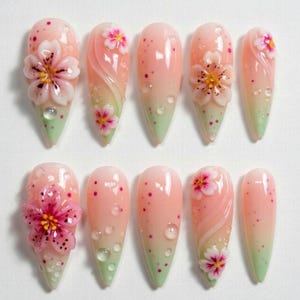 Puede incluir: Un conjunto de diez uñas artificiales con un diseño degradado. Las uñas hacen la transición de un rosa claro a una punta verde. Cada uña presenta un diseño floral en 3D con pétalos rosas y centros amarillos, y algunas tienen detalles de gotas de agua.
