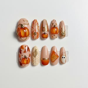 May include: A set of ten press-on nails with autumn-themed designs. The nails feature pumpkins, leaves, and a ghost, with colors including orange, gold, and beige. Some nails have 3D elements and glitter accents.