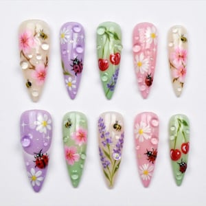 Puede incluir: Un conjunto de diez uñas postizas en forma de almendra, cada una decorada de forma única con diseños florales e insectos. Las uñas presentan fondos rosas, morados y verdes con flores pintadas, mariquitas, abejas y gotas de agua.