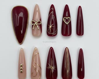 May include: A set of ten artificial nails in various designs. The nails feature a mix of burgundy, nude, and gold accents, including a bow, heart, and starburst patterns. The brand name "SeraphineDesignsCo" is visible.
