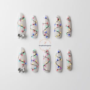 Peut inclure: Dix faux ongles en forme d'amande, peints en gris clair avec un motif de guirlande lumineuse de Noël colorée noire. Deux ongles ont des nœuds argentés et un a un flocon de neige doré. La marque "SeraphineDesignsCo" est au centre.