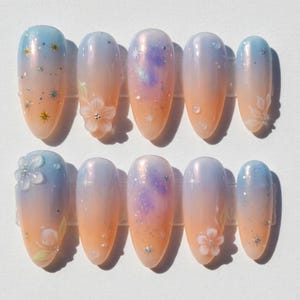 Puede incluir: Juego de diez uñas postizas ovaladas. Cada uña presenta un diseño degradado en azul, naranja y blanco, con purpurina y detalles florales. Algunas uñas tienen adornos florales en 3D y purpurina en forma de estrella.