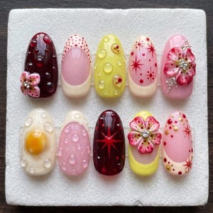 Può includere: Un set di dieci unghie finte con vari design. Le unghie presentano colori come bordeaux, rosa e giallo, con abbellimenti come fiori, pois e motivi a stella. Alcune unghie hanno una finitura lucida, altre un aspetto opaco.
