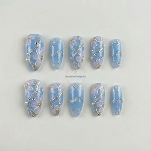 May include: A collection of ten light blue press-on nails. Each nail is decorated with a floral pattern, silver detailing, and tiny pearl accents. The oval-shaped nails have a shiny finish, ideal for a manicure.
