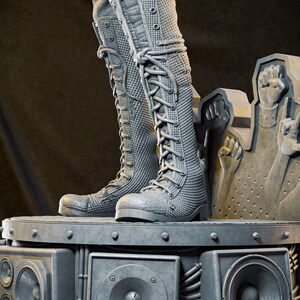 May include: Gray, textured knee-high boots with lace-up closures. The boots are on a platform with speaker components and a sculpture of raised fists. The boots have a detailed, quilted pattern and are made of a durable material.