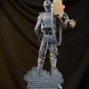 May include: A gray sculpture of a figure with a mohawk and a guitar, standing on a brick wall base. The figure wears a jacket with spikes and holds the guitar over its shoulder. The sculpture is detailed and has a punk rock aesthetic.