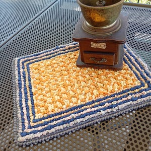 May include: A handmade rag rug with a yellow and orange centre, framed by blue and white stripes. An antique coffee grinder with a wooden base and metal top rests on the rug. The grinder has a small drawer and a label that reads "CAFE".