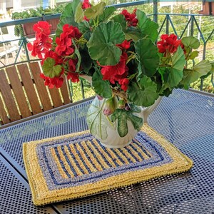 May include: A rectangular, hand-hooked rug with a yellow and blue striped design. The rug has a blue border. A white ceramic jug with fig and leaf patterns holds red flowers and green foliage. The scene is set on a metal table, possibly on a balcony.