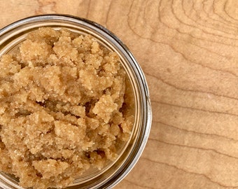 Puede incluir: Un tarro de vidrio transparente lleno de exfoliante de azúcar moreno claro y grueso. El tarro está sobre una superficie de madera marrón claro con vetas visibles. El exfoliante parece granulado y texturizado, llenando el tarro hasta arriba.