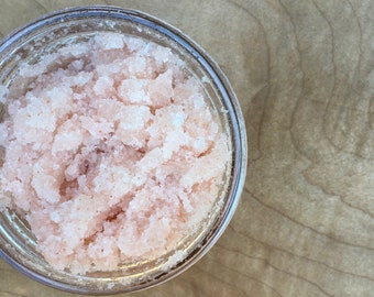 Puede incluir: Una vista de cerca de un frasco de vidrio lleno de un exfoliante de azúcar rosa y blanco. El exfoliante está hecho con cristales de azúcar gruesos y tiene una textura suave y cremosa.