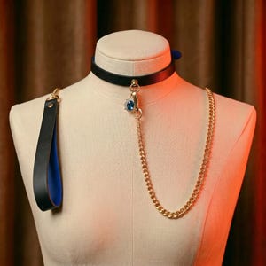 Op de afbeelding: Een zwarte leren halsband met een gouden ketting en een blauwe edelsteen hanger. De halsband heeft een bijpassende zwart-blauwe handgreep. De ketting is met een gouden sluiting aan de halsband bevestigd.