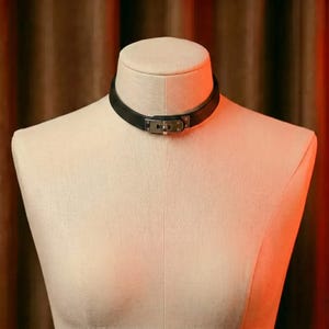 May include: A black leather choker necklace featuring a central silver-toned metal buckle closure. The band is smooth and uniform in width, encircling the neck of a beige fabric mannequin. The hardware includes a rectangular clasp mechanism.