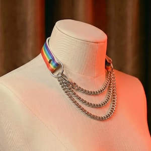 Pode incluir: Um colar gargantilha com listras de arco-íris com três camadas de correntes prateadas. O colar é exibido em um manequim. As cores do arco-íris incluem vermelho, laranja, amarelo, verde, azul e roxo. O colar tem um anel e fecho de metal prateado.