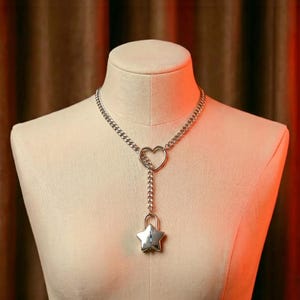 Peut inclure: Un collier argenté avec un pendentif en forme de cœur et un cadenas en forme d'étoile. Le collier est présenté sur un mannequin. La chaîne est composée de petits maillons entrelacés.
