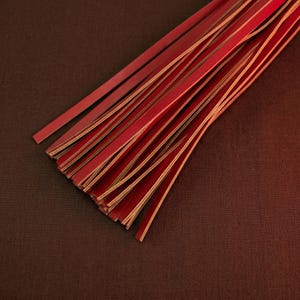 May include: A collection of thin, red leather strips with a gold-coloured edge. The strips are arranged in a fan shape, with some overlapping. The background is a dark brown, textured surface.