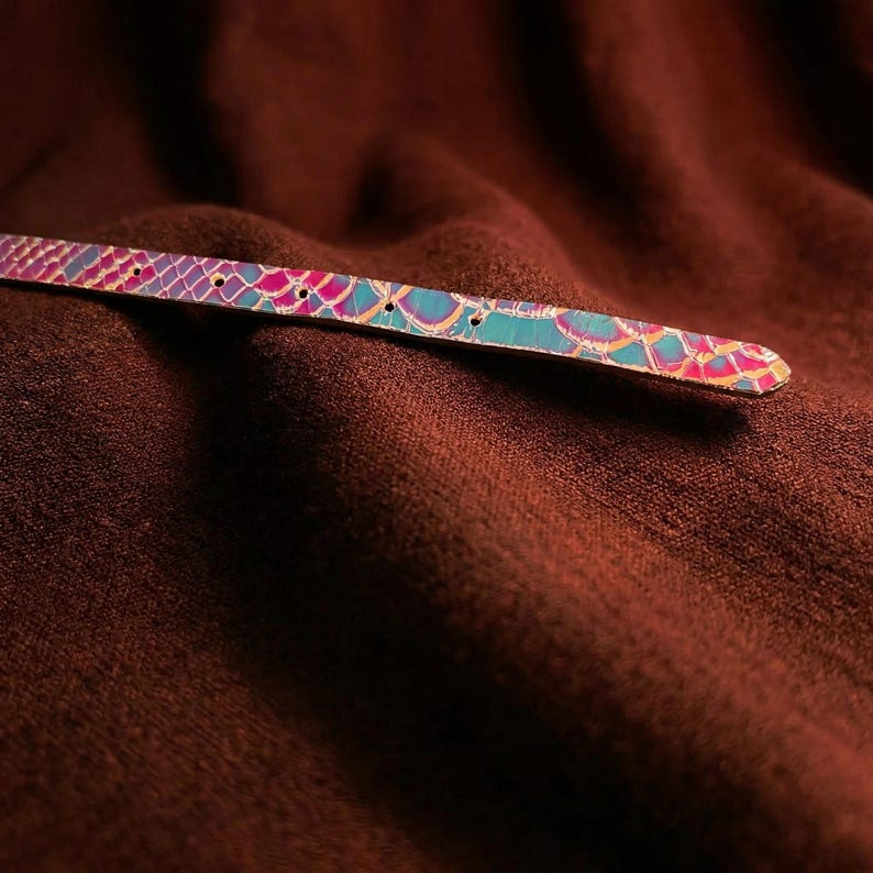 May include: A colorful, iridescent belt with a scale-like pattern in shades of pink, blue, and purple. The belt has several holes for adjustment and is set against a dark brown, textured background.