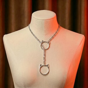 May include: Silver-toned cat-shaped pendant necklace. The necklace features a chain design with two cat head pendants, one at the top and one at the bottom, connected by a chain. The necklace is displayed on a mannequin.