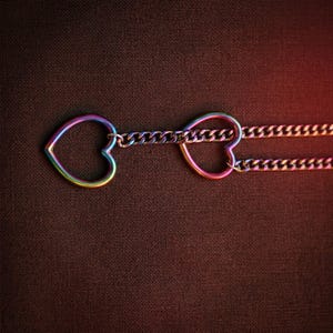 Puede incluir: Un collar con dos dijes en forma de coraz&oacute;n abierto conectados por una cadena de eslabones. El metal tiene un acabado iridiscente de colores del arco&iacute;ris que cambia entre tonos de azul, verde, amarillo y rosa sobre la superficie.