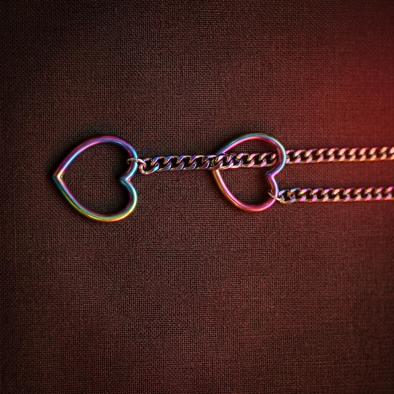 Puede incluir: Un collar con dos dijes en forma de coraz&oacute;n abierto conectados por una cadena de eslabones. El metal tiene un acabado iridiscente de colores del arco&iacute;ris que cambia entre tonos de azul, verde, amarillo y rosa sobre la superficie.
