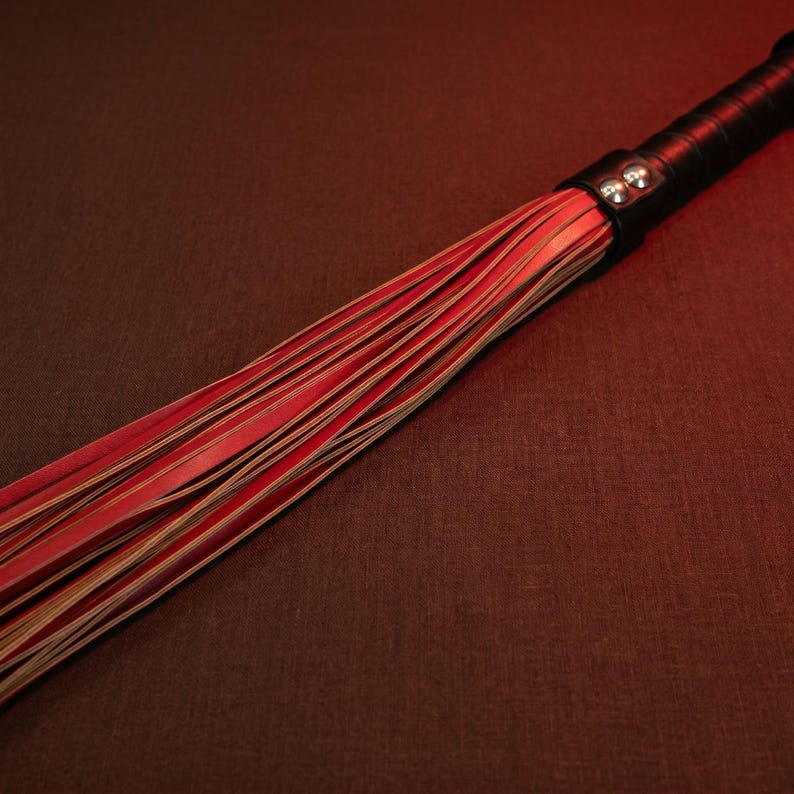 May include: A red and tan leather flogger with a black handle. The handle is wrapped in black leather and secured with a silver-toned metal band. The flogger's strands are a vibrant red.