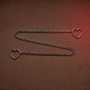 Puede incluir: Una cadena de metal con un acabado iridiscente de colores del arco&iacute;ris. La cadena est&aacute; dispuesta en un patr&oacute;n de zigzag y presenta un anillo en forma de coraz&oacute;n en cada extremo. El metal tiene un aspecto reflectante y multicolor entre azul y oro.