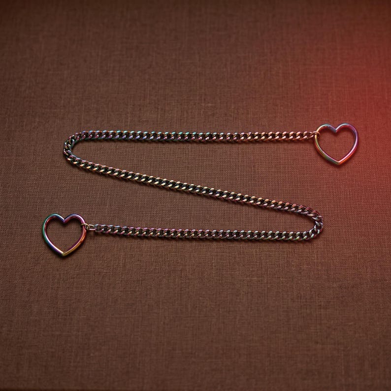 Puede incluir: Una cadena de metal con un acabado iridiscente de colores del arco&iacute;ris. La cadena est&aacute; dispuesta en un patr&oacute;n de zigzag y presenta un anillo en forma de coraz&oacute;n en cada extremo. El metal tiene un aspecto reflectante y multicolor entre azul y oro.