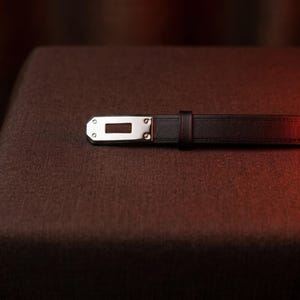 May include: A dark brown leather belt featuring a polished silver-tone metal buckle with a rectangular cutout and two small rivets. The belt strap has visible stitching along the edges and includes a matching dark brown leather keeper loop.