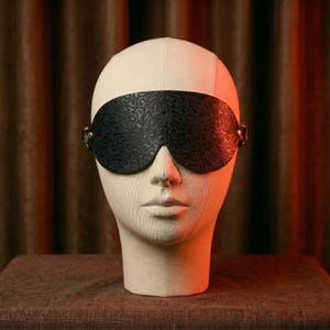 May include: Black leather eye mask with an embossed floral pattern. The mask has a curved shape to fit the face and is secured with small gold-colored buckles on each side. The mask is displayed on a mannequin head.