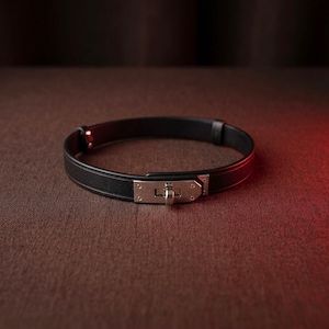 May include: A black leather bracelet featuring a silver-toned metal clasp mechanism. The band is smooth and circular, with a polished metallic closure centered on the front. The design includes small rivets securing the strap to the hardware.