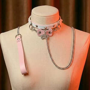 Könnte beinhalten: Ein weißes und rosa Halsband mit passender Leine. Das Halsband hat silberfarbene Hardware, herzförmige Akzente und ein rosa schleifenförmiges Mittelstück. Eine silberne Kette erstreckt sich von der Schleife. Die Leine ist einfarbig rosa.