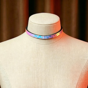 May include: A choker necklace with a black band and a holographic, iridescent design. The choker features a repeating pattern of small, interconnected shapes that reflect a range of colors, including pink, blue, and yellow. The necklace is displayed on a mannequin.