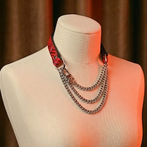 Op de afbeelding: Een rode leren halsband met een zwart bloemenpatroon en zilverkleurige metalen hardware. Drie gelaagde zilverkleurige stoeprandkettingen hangen aan de voorkant, verbonden door een karabijnhaak aan de halsband op een etalagepop.