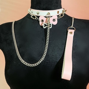 Op de afbeelding: Een witte en roze halsband met een zilveren ketting en riem. De halsband heeft hartvormige uitsparingen en een vlindervormige bedel. De riem is roze met een zilveren hartdetail.