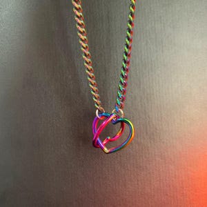Puede incluir: Un collar que presenta una cadena de eslabones con un acabado de arco&iacute;ris iridiscente y dos colgantes entrelazados en forma de coraz&oacute;n. Los eslabones met&aacute;licos y los corazones muestran un espectro de colores azul, verde, amarillo, naranja y violeta.